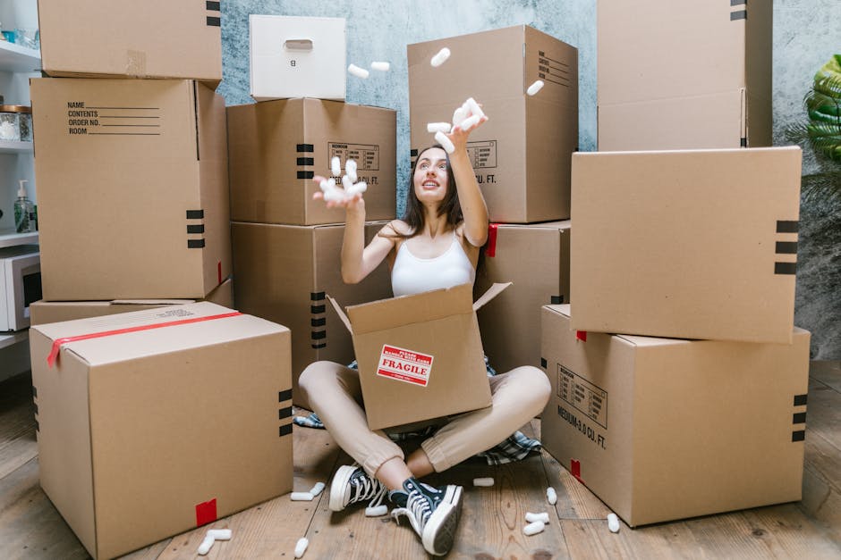 A young woman with long brown hair, sitting cross-legged on a wooden floor inside a room, is surrounded by numerous cardboard boxes of varying sizes, some with labels and packing tape. She is wearing a white sleeveless top, beige pants, and black-and-white sneakers. The woman is smiling and tossing small white packing peanuts into the air, while an open box with a red 'Fragile' sticker sits on her lap. Behind her, additional packed boxes are stacked against a textured light blue wall, and the room appears to be prepared for home relocation or furniture transport as part of a house move. A partial view of a work surface with a potted plant and other packing materials can be seen to the left. The scene captures the packing process associated with professional removals services provided by Man with Van Hammersmith, demonstrating packing and moving activities typical of house removals on Fulham Palace Road.
