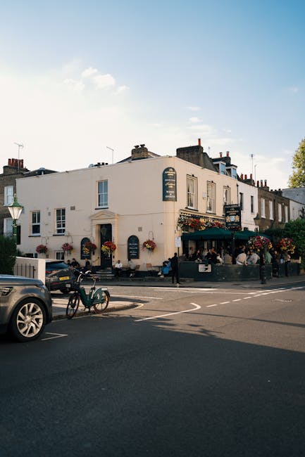 An exterior view of a traditional Pub located on Fulham Palace Road during daytime, showing a white building with multiple windows and flower boxes on the ledges. The pub has outdoor seating with dark green awnings and tables, some with people seated. In front of the building, vehicles including a black car, a bicycle, and a scooter are parked or passing by on the street. The scene is illuminated by natural daylight with a clear sky overhead. This setting represents a typical urban environment suitable for home relocation and furniture transport services offered by companies like Man with Van Hammersmith, as they assist with house removals, packing, and logistics in the Hammersmith area.