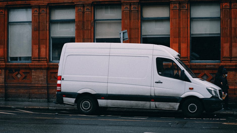 A silver panel van parked on a city street in front of a brick building with large stained glass windows and a stone entrance. The van is positioned close to the curb, with its front portion visible i