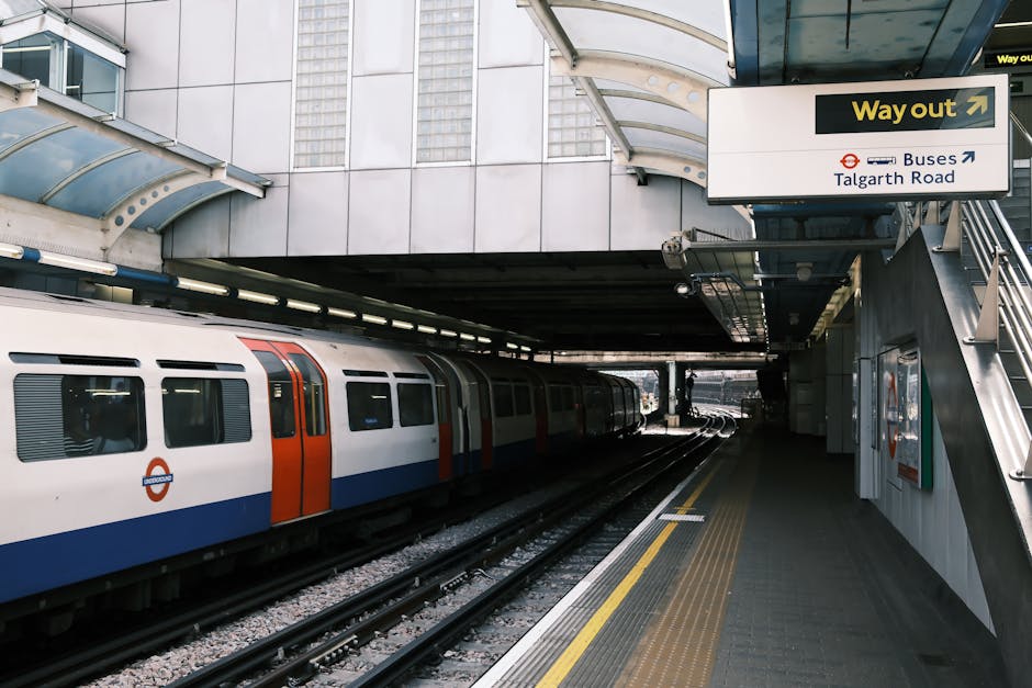 The image shows a modern London underground train with a curved white body accented by an orange and blue stripe, stationed at an outdoor platform of an urban train station, possibly in the Hammersmith area. The platform features yellow tactile paving along the edge for safety, and has a grey tiled surface. Overhead, there is a clear, curved canopy supported by metal beams, providing shelter. A prominent white directional sign is mounted above, indicating exits towards 'Way out' with an arrow, and listing nearby destinations including 'Buses' and 'Talgat Road.' To the right, metal staircases lead upwards, and the station infrastructure includes wall-mounted safety notices and equipment, used during the process of home relocation or furniture transport logistics. The scene is illuminated by natural daylight, with shadows cast in the shaded areas, emphasizing the urban transit environment connected to house removals and moving services provided by Man with Van Hammersmith.