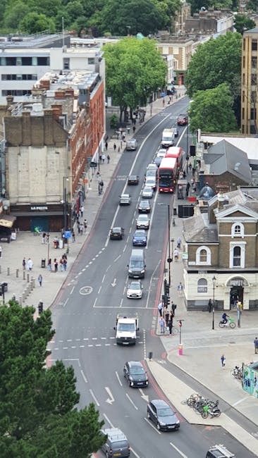 An aerial view of a busy city street during daytime showing a traffic lane with multiple vehicles including cars, vans, and buses moving along a slightly curved road, with pedestrians walking on the wide sidewalks. On the left side, there are brick and modern commercial buildings, while on the right side, there are residential houses and a small shop with a sign. Several large, leafy green trees line the street, providing shade and separation between the traffic and pedestrian areas. In the foreground, a silver van belonging to Man with Van Hammersmith is parked near the curb, possibly involved in home relocation or furniture transport services. The scene captures an active urban environment consistent with house removals and packing and moving logistics, with the environment well-lit by natural daylight.