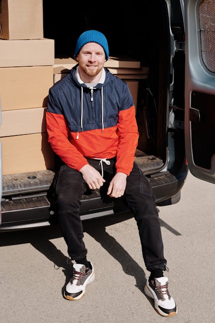 A man wearing a blue beanie, a navy and red windbreaker jacket, and black pants is seated on the edge of the open rear door of a black moving van. Inside the van, several large cardboard boxes are stacked neatly, some wrapped in plastic or protective packing materials. Additional smaller boxes and packing supplies are visible, indicating an organized relocation process. The man is smiling, with his hands resting on his thighs, and appears ready to assist with furniture transport during a home relocation. The surrounding environment shows an outdoor setting, with the van parked on a paved surface, and natural daylight illuminating the scene. The image reflects the loading process involved in professional removals services provided by Man with Van Hammersmith, emphasizing careful packing and transport of household items.