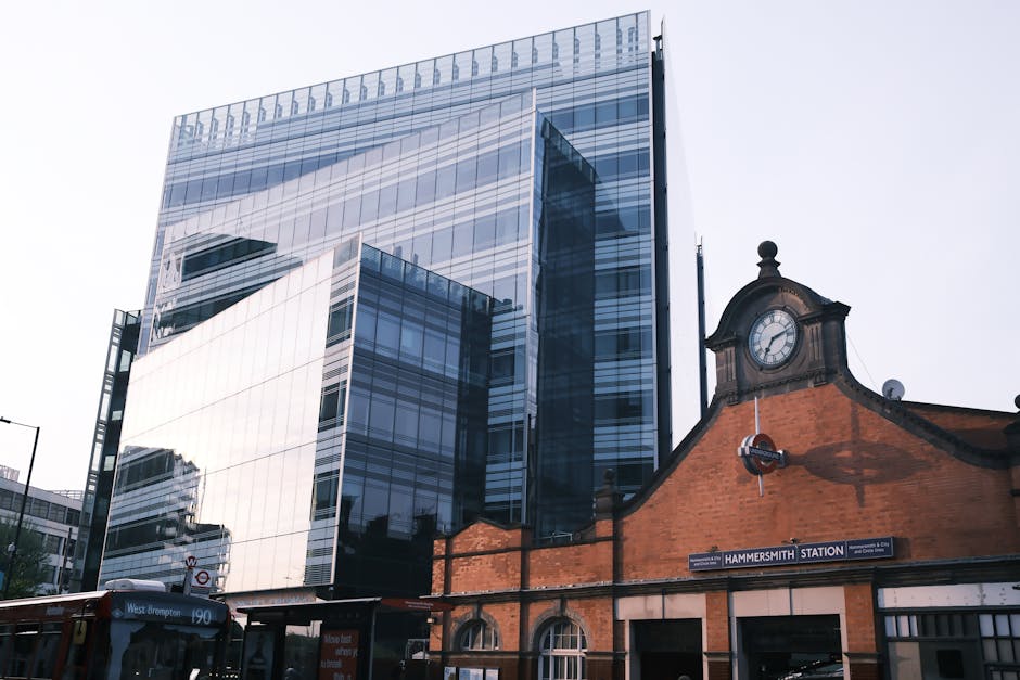 A modern, multi-storey glass building with reflective windows towering above a historic brick railway station structure at Hammersmith station. The station features a traditional clock face set within an ornate roofline, with a sign reading 'HAMMERSMITH STATION' attached to the brick facade. In the foreground, part of a bus with the route number 190 is visible on the street, along with a glimpse of pavement and some street signage. The area is well-lit with natural daylight, showing the contrast between the contemporary commercial building and the older station architecture. This image captures the ongoing home relocation process, where furniture and packing materials might be moved through the station, with [COMPANY_NAME] performing loading or unloading activities involving boxes, furniture, or moving equipment within this urban environment, as part of professional removals services provided in Hammersmith.
