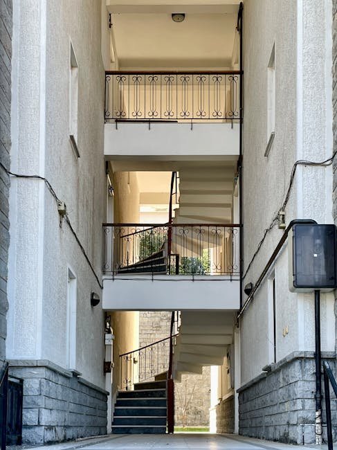 The image shows the interior of a multi-storey residential building with three floors, featuring a central stairwell with white-painted concrete stairs and black metal railings. Each floor has a small balcony with decorative black wrought iron railings and some potted plants visible on the middle balcony. The walls are light-colored with small rectangular windows on each level. The stairwell is lit by natural daylight coming from an open entrance at the ground level, where a pavement and a step lead into the building. Some electrical conduit wiring runs along the walls, and a security camera is mounted near the ceiling at the top of the stairwell. This setting is typical of apartment blocks used for house removals, relocation logistics, or moving services, as companies like Man with Van Hammersmith facilitate furniture transport and packing during home relocations.