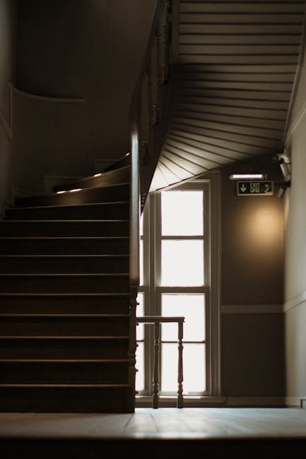 An interior stairwell in a residential building with a curved wooden staircase featuring dark wooden steps and a polished wooden handrail. The stairs ascend to an upper floor, with a large window at the landing allowing natural light to illuminate the area. Adjacent to the staircase is a door with a vertical window panel, leading to an outside space or corridor. On the wall near the door, there is a green illuminated exit sign indicating the emergency escape route. The scene captures the clean, well-maintained environment typical in a home or apartment building during a home relocation process, with the natural lighting highlighting the structural features. This setting aligns with the context of house removals and furniture transport, as experienced by professional moving services such as Man with Van Hammersmith.