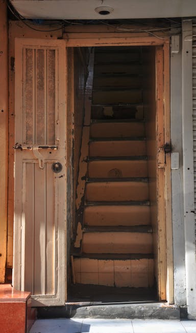 The image shows the interior of a multi-storey residential building with three floors, featuring a central stairwell with white-painted concrete stairs and black metal railings. Each floor has a small balcony with decorative black wrought iron railings and some potted plants visible on the middle balcony. The walls are light-colored with small rectangular windows on each level. The stairwell is lit by natural daylight coming from an open entrance at the ground level, where a pavement and a step lead into the building. Some electrical conduit wiring runs along the walls, and a security camera is mounted near the ceiling at the top of the stairwell. This setting is typical of apartment blocks used for house removals, relocation logistics, or moving services, as companies like Man with Van Hammersmith facilitate furniture transport and packing during home relocations.