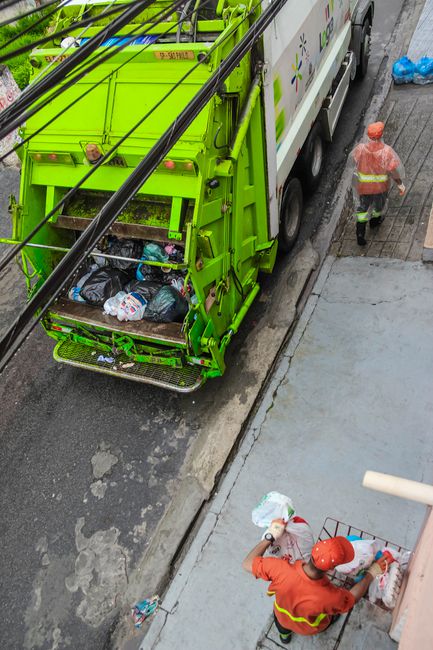 A row of four large white recycling bins with lids, arranged side by side on a paved surface outside a property, with labels indicating they are for waste disposal. To the left, there is a dark bin ma